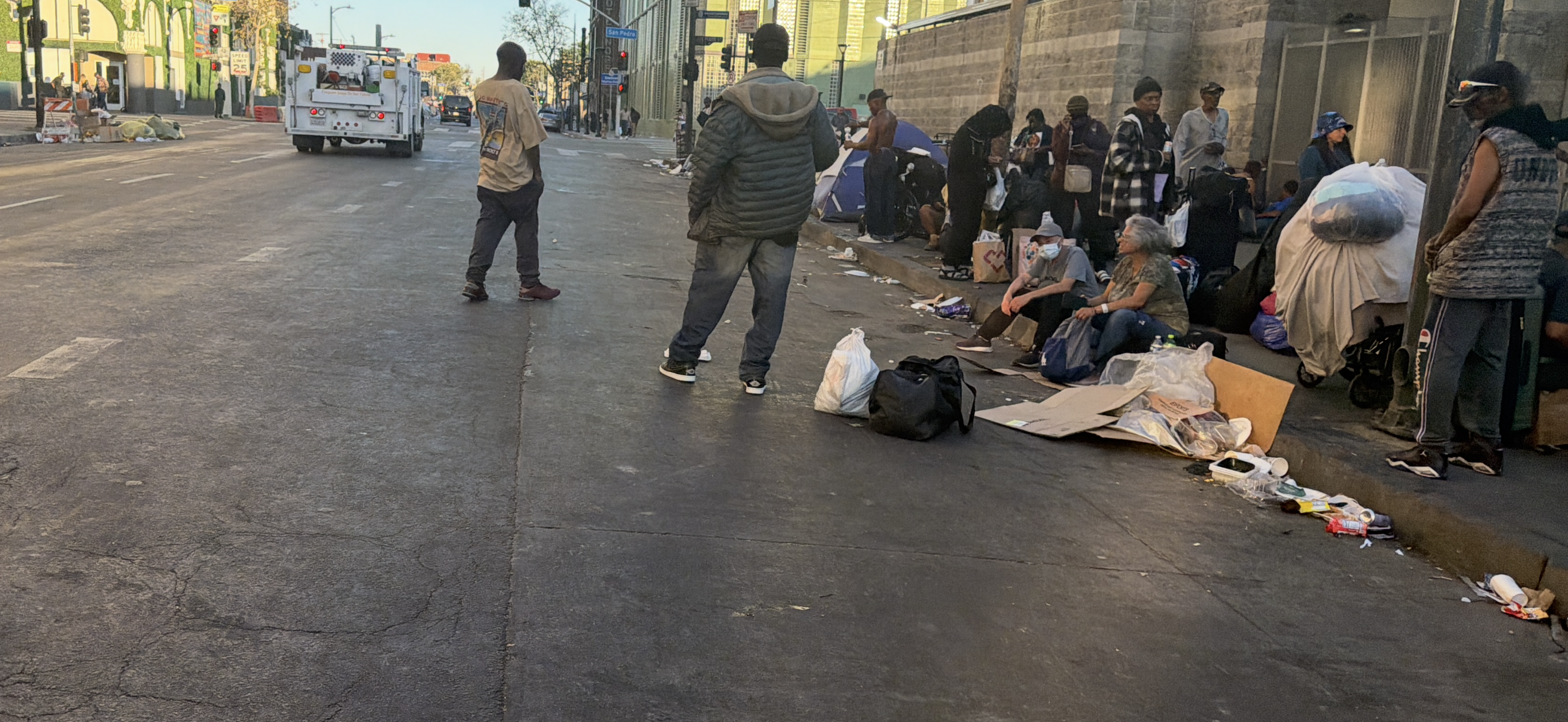 Unhoused residents gather along a sidewalk in Skid Row as outreach and sanitation activity unfolds nearby in downtown Los Angeles.