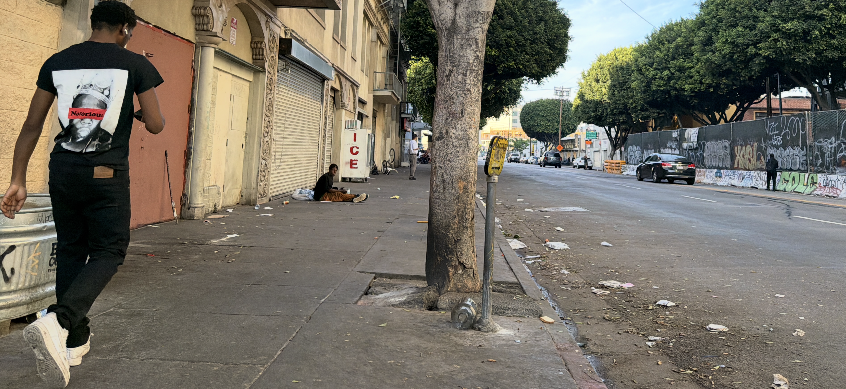 A pedestrian walks along a downtown Los Angeles sidewalk while another person sits near a storefront, Saturday, March 2026. Advocates say unstable living conditions can make it harder for unhoused individuals to receive continuous care as serious illness progresses.